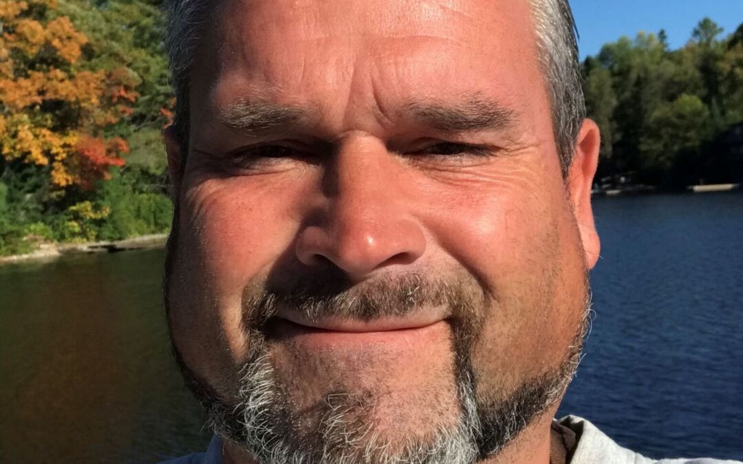 a selfie of Danny with a beautiful Ontario lake and trees behind him.